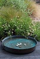 A broad and shallow copper bowl creates a simple water feature on the edge of a wooden deck, beside a clump of fleabane.