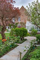 A path leads through a small formal kitchen garden with Malus domestica 'Falstaff' - Apple - in blossom, trained along a stepover cordon edging mixed beds, view of period house beyond 