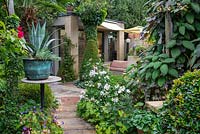 Path leads to a garden room, passing an Agave americana perched on a table, and edged in begonias, hardy geraniums, Japanese anemones, dahlias and phlox.