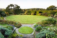 View over circular lawns edged with paving and gravel, surrounded by borders of shrubs and herbaceous perennials, with formal hedge beyond