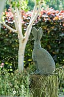 Sculpture of a hare on a wooden plinth in front of Betula utilis var. jacquemontii 'Grayswood Ghost' and a purple beech hedge. Stevington Manor Garden