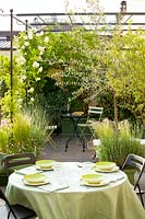 Terrace with bistro set laid for lunch with mixed planting including rose 'brise parfum'