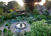 Overview of garden with rooms separated by yew hedges. Paving slab path between borders of shrubs and seating area beside water feature fountain. 