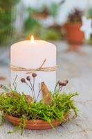 White candle on terracotta plate decorated with moss, dried lime foliage and string
