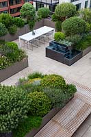 Looking down on city roof garden with water feature and seating area surrounded by perimeter of troughs filled with evergreen shrubs