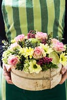 Person holding a floral display, pot wrapped with birch bark with Rosa 'Cupcake' - Rose, Chrysanthemum 'Alero Cream', Dianthus 'Green Trick', Hydrangea 'Groen' and pink Pepperberries and foliage