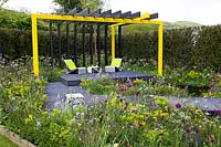 Black and yellow steel structure above black seating area and wooden platform boardwalk, surrounded by naturalistic meadow style planting in 'Urban Oasis' at RHS Malvern Spring Festival 2018