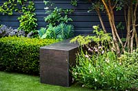 Concrete cube with porcelain cladding finished in a dark steel effect surrounded by with Gaura lindheimeriÂ 'Sparkle White', Osmanthus x burkwoodii, Taxus baccata and metal cube by black wooden fence. 