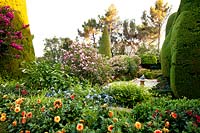 View over flower beds to paved terrace with shrubs, large topiary forms and trees beyond