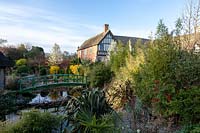 View across foliage bed to pond with Monet bridge, views of shrubs and trees around half-timbered house 
