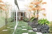 Enclosed terrace with paving stones amongst Ophiopogon japonicus 'Nanus' groundcover and a bed of shrubs and perennials including Ophipogon 'Nigrescens', Iris japonica and Acer palmatum