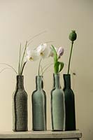Fabric bottles containing Orchid, Tulip and poppy seed head on shelf. 