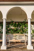 View under arch of a loggia with wall-trained Rosa - Rose - behind wooden bench 