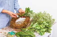 Woman using floristry wire to fix bundle of cut flowers and foliage to moss wreath form. 