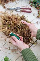 Woman using moss, attached with floristry wire, to build up wreath form around wire frame