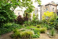 Square garden with tall buildings all around, wooden benches, paths and mixed planting including trees, ornamental grasses and shrubs 