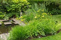 An informal suburban country garden with perennials, such as Hemerocallis, in a bed near small pond with paving and stepping stones 