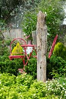 A freestanding rustic timber branch with a plastic rain gauge mounted on in front of red outdoor table and chairs.