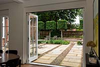 View from inside house through open doors to patio with bands of pavers and bricks to clipped Carpinus - Hornbeam - square trained trees