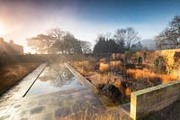 Contemporary garden within an old walled garden, with long pond in paved area with block planting of ornamental grasses with dead stems and seedheads 