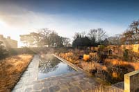 Contemporary garden in old wall garden, view across paved area with long rectangular pond with block planting of ornamental grasses and other shrubs