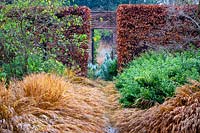 View over double border of ornamental grasses, evergreen shrubs and trees to a clipped hedge with view through 