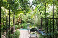 View through metal archway to tropical-style urban garden, tiled dining terrace with a mix of palms, ferns and other foliage plants 