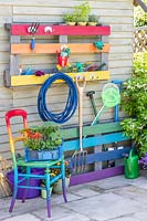 Rainbow coloured pallet tool organiser attached to outside wall of a shed with matching chair 