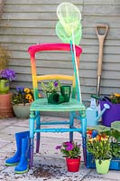 Wooden chair painted in rainbow colours on patio with bedding plants, blue welly boots and children's fishing nets