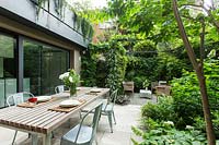Garden outside house divided into a dining area and more relaxed seating area by metal pergola and climbers, perimeter of property screened by foliage trees and shrubs