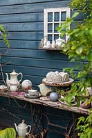 A collection of eclectic objects: tea cups, coffee pots, a cake plate and sauce boat, displayed on old timber shelf near veranda wall