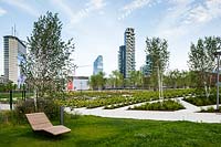 Public park with grass, seating and young trees with beds and paths, cityscape beyond 