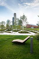 Public park with seating on grass and recent plantings of young trees, geometric beds with paths 