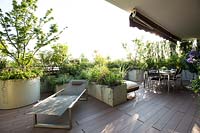 View out to the open areas of the decked terrace, a sun lounger near container with Acer palmatum 'Crispifolium' and other planters with mixed foliage. Unrolled shade canopy attached under ceiling and other seating options beyond.
