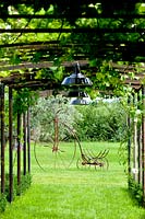 View under pergola covered with Vitis - Vine, grass path and overhead lamps. Vintage bike on lawn beyond.