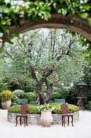 View through archway to seating area by circular raised bed with central tree.