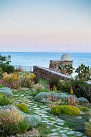 View over terrace wall to the sea. Planted with drought tolerant and wind resistant planting, mixing Mediterranean shrubs: Pistachia lentiscus, Myrtus communis, Helichrysum with plants from similar climates:  This Aloe and Agave, Stipa tenuissima, Muhlenbergia capillaris, Chondropetalum tectorum and Tamnochortus insignis