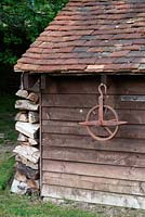 Old wooden shed with tiles roof, log pile and an old pulley wheel hanging from the roof