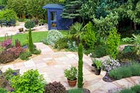 View over paved area to the blue painted garden room with curved roof. Plants in beds: Trachycarpus fortunei - Palm, Eriobotrya japonica, Tetrapanax papyrifer, Lavandula - Lavender, variegated Pittosporum, pencil conifers and Hebes 