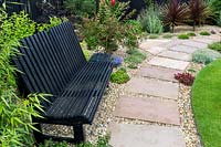 A wide, paving and gravel pathway near a black slatted bench