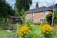View of flint cottage and garden, featuring Rudbeckia fulgida var. sullivantii 'Goldsturm', benches and wooden shed