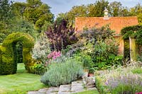 Looking across the garden in the early sunlight to the Clock House, via the yew hedge and its keyhole archways. Plants include Lavandula x intermedia, phlox, Elaeagnus 'Quicksilver', Cotinus coggygria and Rosa glauca.