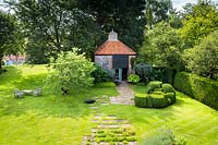 View over lawn with mosaic of paving stones leading to Buxus - Box- topiary and 18th century dovecote, also in lawn is a Cercis Siliquastrum - Judas Tree - and seating