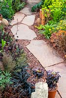 Stone path with granite chips, alongside border of mixed plants and garden bric-a-brac - RHS Malvern Spring Festival. 