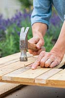 Woman using nails and hammer to fix pieces of narrow wood strips to fill gaps in pallet. 