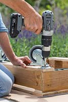 Woman using electric screwdriver to fix castors on to bottom of pallet. 