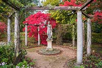 Classical statue framed by pergola with backdrop of red from Acer palmatum 'Bloodgood' - Japanese Maple