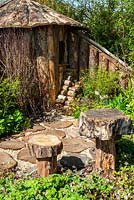 Path of tree trunk slices and wood chips, leading to rustic wooden hut with logs alongside and structural features in border  - RHS Malvern Spring Festival