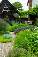 Assorted herbs on gravel bed, including Bronze Fennel, Nepeta, Santolina, Golden Marjoram, Purple Sage, Cotton Lavender, Phlomis and Thyme with bird feeders on end of barn and table and chairs beneath Wisteria - Open Gardens Day, Coddenham, Suffolk