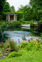 Garden pond with border of Primulas and Astilbes, small  fountain and dinghy moored alongside jetty. Summerhouse on far bank - Open Gardens Day, Palgrave, Suffolk